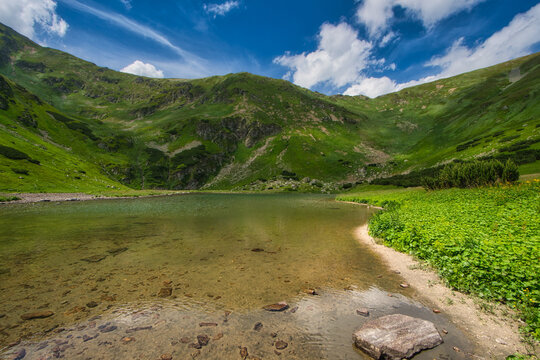 View of a serene tarn mirroring the vibrant green slopes under a sky streaked with clouds, a picturesque haven of nature's artistry, Rackove pleso, Zilina Region, Slovakia.