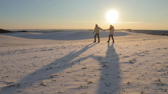 Young couple walks casting long shadows on snow at winter sunset. Romantic man and woman join hands during promenade on frozen valley. Happy tourists enjoy cold dusk in nature