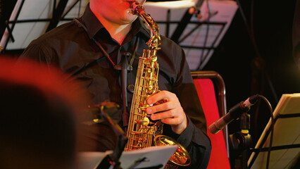 Obraz premium A focused musician plays a golden saxophone in an orchestra. Mid-shot with warm, red stage lighting highlighting the instrument and player, creating a sophisticated and professional mood