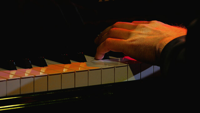 A close-up shot of a musician's hand playing a grand piano. Dramatic, warm lighting highlights the keys and creates an intimate, focused mood. Dark background emphasizes the performance