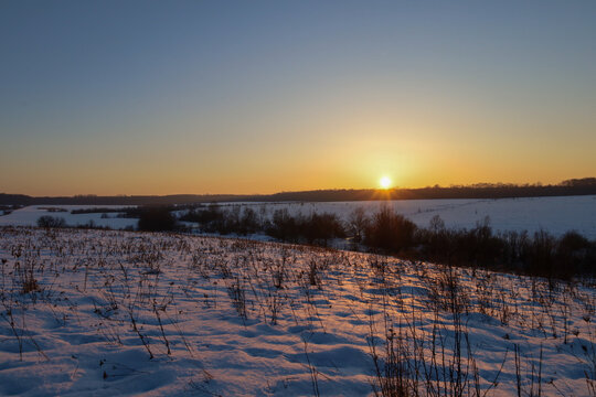 Scenic winter landscape featuring a panoramic view of snow-covered hills and fields during a golden sunset. Bright sun rays illuminate the sparkling snow and dry grass, creating an atmosphere of peace