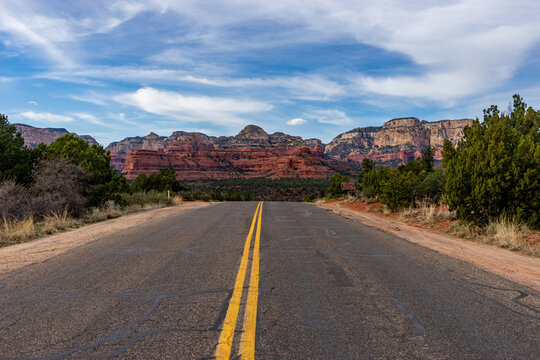View of a long, straight asphalt road with double yellow lines slicing through the arid landscape towards towering red rock formations, Winslow, Arizona, United States.