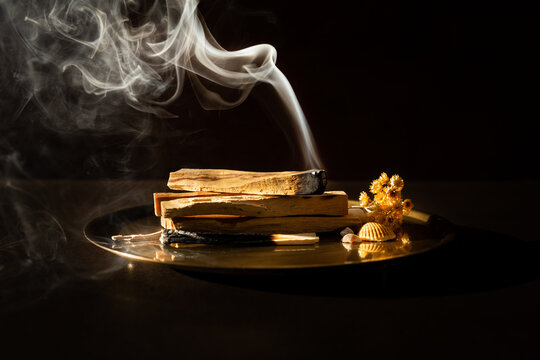 Un b&acirc;ton de Palo Santo allum&eacute; pos&eacute; sur une pile de b&acirc;tons sur un plateau en bronze, vue de pr&egrave;s et de face, belle fum&eacute;e blanche qui ondule sur fond brun fonc&eacute;
