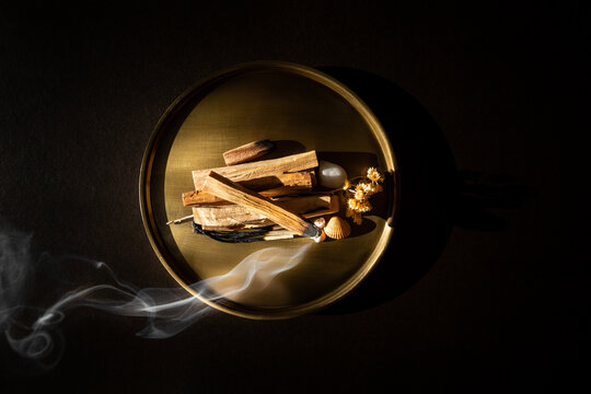 Palo Santo pos&eacute;s sur un plateau en bronze, vue du dessus, un b&acirc;ton allum&eacute; sur le haut de la pile se consumant, fum&eacute;e blanche qui s'&eacute;tire et ondule vers la gauche, ambiance douce et m&eacute;ditative