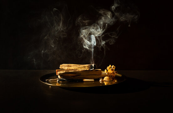 B&acirc;tons de Palo Santo en fagot pos&eacute;s sur un plateau en bronze, un b&acirc;ton sur le dessus br&ucirc;lant et lib&eacute;rant une fum&eacute;e blanche verticale, fond brun fonc&eacute;, ambiance m&eacute;ditative et douce