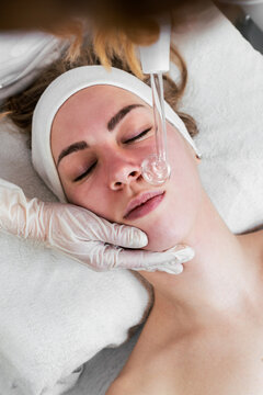A professional beautician performing a high frequency darsonvalization procedure on a young woman's face to improve skin health and treat acne in a modern spa clinic.