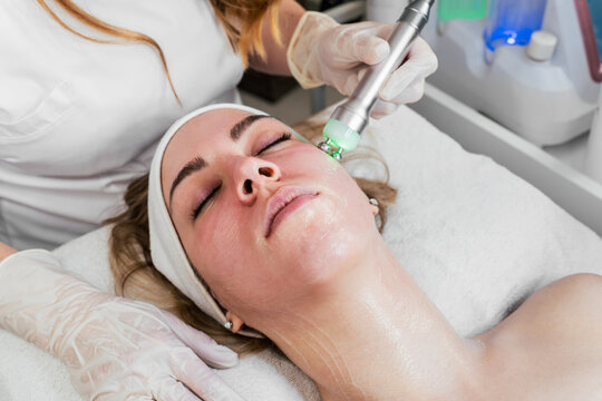 Professional esthetician performing a microcurrent miolift facial treatment on a young woman using a device with green LED light therapy for skin rejuvenation and anti-aging in a spa.