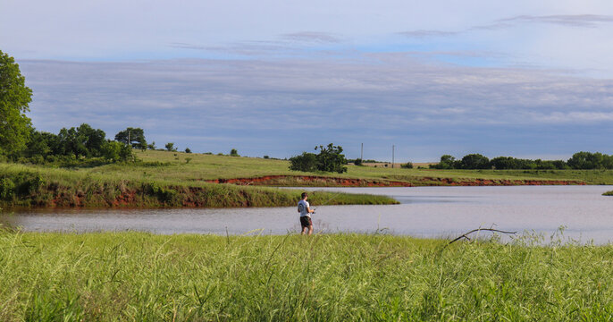 View of a lone fisherman casts his line into the tranquil lake, framed by the vibrant green of tall grass and the earthy red bluffs, Oklahoma City, Oklahoma, United States.
