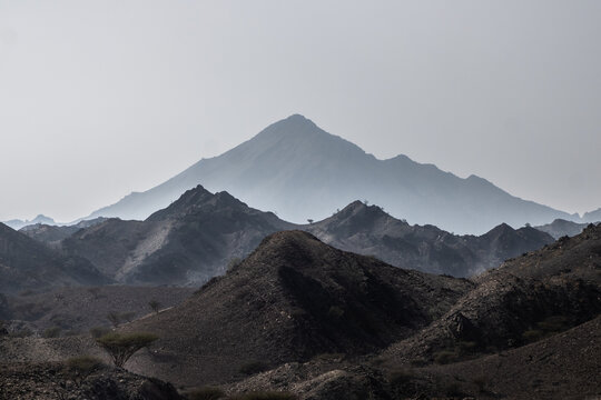 View of stark, rugged mountains under a hazy sky, casting long shadows across the arid landscape, Wadi Bani Khalid, Ash Sharqiyah North Governorate, Oman.