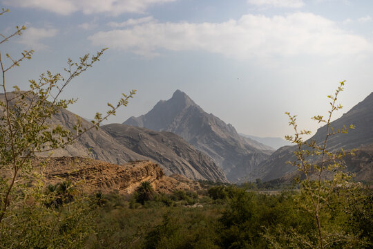 View of rugged, sun-kissed mountains rise majestically against a hazy sky, framing a lush valley teeming with verdant foliage, Wadi Bani Khalid, Oman.