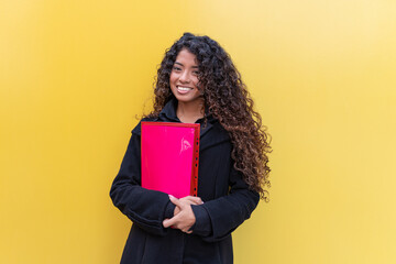 Fototapeta premium Young woman student holding a pink binder, smiling and looking at camera against a vibrant yellow background