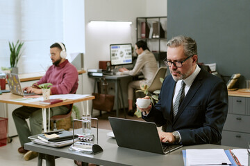 Obraz premium Middle aged Caucasian man wearing glasses working on laptop and drinking coffee in modern office, young man with headphones and woman in suit working at desks in background