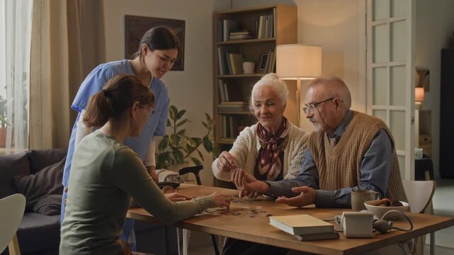 Friendly female nurse in medical scrubs helping group of seniors at table with puzzles in cozy retirement home