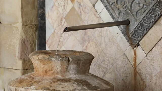 Ornate metal spout pours mineral water into a stone vase against a textured marble tile wall in a historic spa setting, showcasing natural thermal spring source details.

