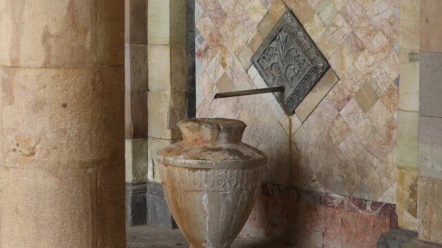 Ornate metal spout pours mineral water into a stone vase against a textured marble tile wall in a historic spa setting, showcasing natural thermal spring source details.

