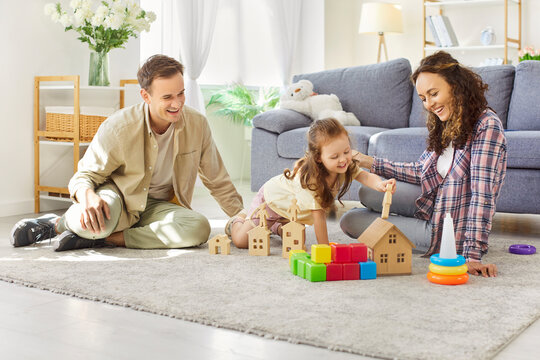 Happy parents and child daughter play at home in educational game with toys. In livingroom, mother and father create wooden houses, laughing together with love. Family play, leisure and learning.