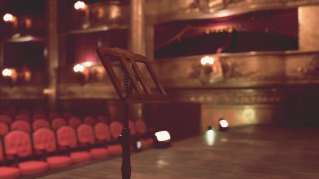 In an ornate old opera house, a music stand stands in the foreground, while rows of red velvet seats lead to an empty stage adorned with rich decorations
