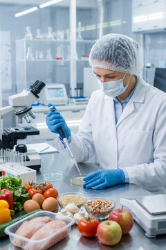 Female food scientist in white lab coat, gloves,mask and hair net using pipette to analyze food samples in modern lab. Professional researcher conducting quality control tests with fresh produce.