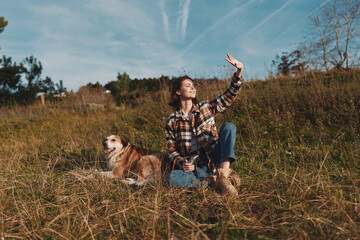 Obraz premium Woman in plaid shirt sits in a grassy field with a dog beside her, raising her arm toward the bright sky during a sunny outdoor moment in nature today.