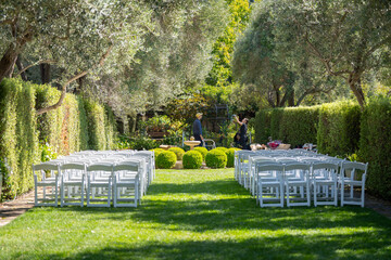 Wedding arch with empty chairs set up in a garden, prepared for an outdoor ceremony. Elegant floral decorations and a serene atmosphere create a beautiful setting for a wedding celebration