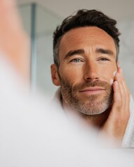 Obraz premium Male applying skincare product to his face while looking in a bathroom mirror, modern bathroom decor with soft lighting and neutral colors visible in the background