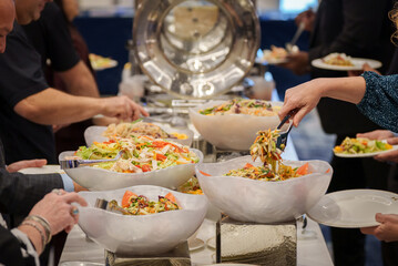 People serving themselves food with tongs from plates at a buffet table. Hands selecting a variety of dishes in a self-service dining setup, typical for events, parties, or restaurants
