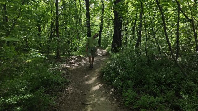 Man walks calmly along narrow forest path surrounded by green trees and summer foliage during quiet outdoor walk in natural woodland environment