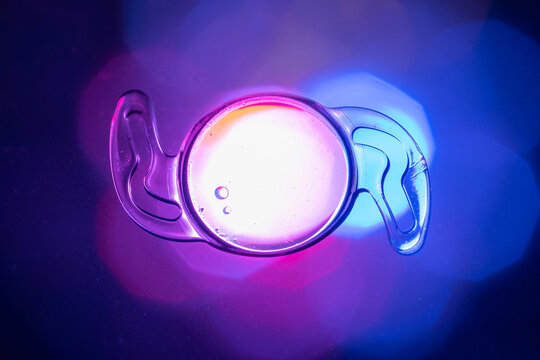 Macro Shot Of A Flexible Intraocular Lens (IOL) With Haptics On A Neon Blue And Purple Backgr