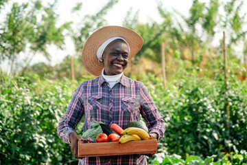 Happy senior Black woman farmer holding a wooden crate of fresh organic vegetables and fruits in a...