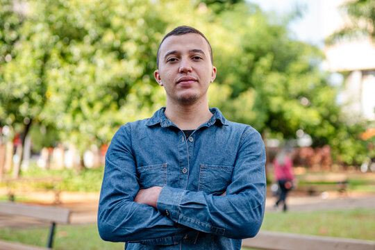 Confident young man in blue denim shirt standing with arms crossed in a public park.