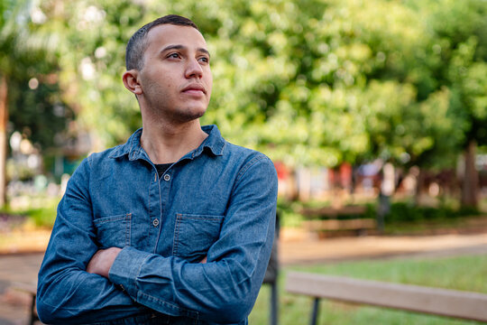 Thoughtful young man in denim shirt standing with arms crossed in a sunny outdoor park setting.