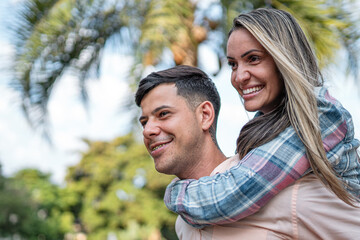 Fototapeta premium Happy young man giving a piggyback ride to his girlfriend in a tropical park, smiling couple enjoying a romantic moment outdoors.