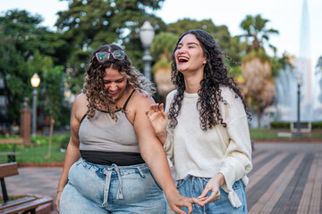 Two diverse female friends with curly hair laughing while walking in a city park during the day.