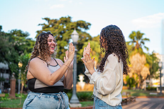 Two happy young women with curly hair laughing and playing a hand-clapping game in a sunny public park