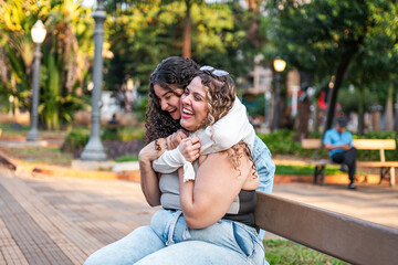 Naklejka premium Two happy female friends embracing and laughing while sitting on a park bench in a sunny public garden.