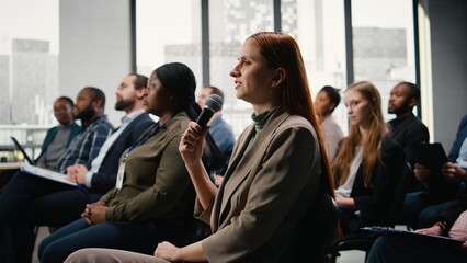 Female participant raising hand and engaging in debate on microphone, attending corporate summit in conference hall with chairs and active audience. Diverse group at professional event.