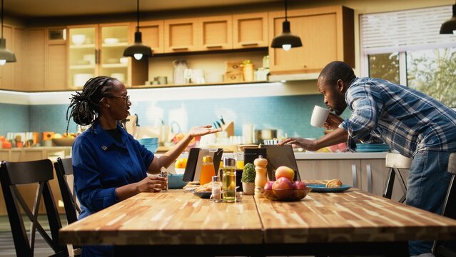 African american man in a hurry running late for his office job, skipping breakfast and leaving his housewife to eat alone at the table. Boyfriend waving goodbye and rushing to leave.