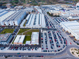 Large industrial area with numerous parked cars and buildings at midday in an urban setting surrounded by fields and roads