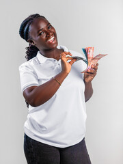 A smiling Black woman in a white polo shirt happily fans out a stack of Ghana cedi banknotes. She has braided hair and poses against a plain grey background, radiating success and financial joy.