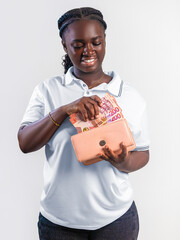 A smiling young woman in a white polo shirt holds a peach-colored wallet, happily pulling out several Ghanaian Cedi banknotes. She has braided hair and stands against a plain white background.