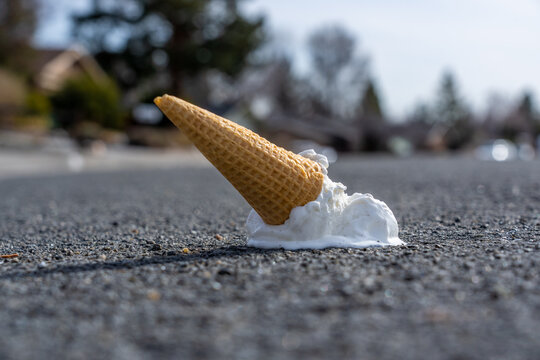 Melting ice cream cone lies on a dark asphalt surface. The beige waffle cone is inverted with white ice cream partially melted around its base, creating a small puddle.