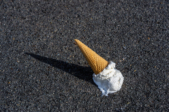 Melting ice cream cone lies on a dark asphalt surface. The beige waffle cone is inverted with white ice cream partially melted around its base, creating a small puddle.