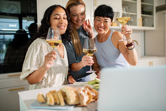 Three women toasting wine during video call in a home kitchen