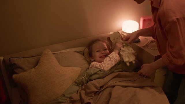 Mother giving plush rabbit to daughter and kissing her cheek while father opening book, preparing to read it