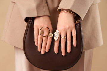 Young woman in beautiful golden rings and bracelet with stylish bag on brown background, closeup