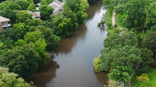 Drone footage shows swollen river overflowing its banks near homes and a park pathway. High water levels indicate flooding conditions affecting a suburban area