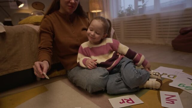 Tilt shot of smiling mother sitting on carpet and hugging her loving daughter while they learning alphabet, using flash cards