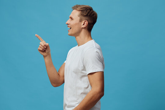 Smiling man pointing finger sideways in profile view on bright blue background, casual white t-shirt, joyful facial expression, studio portrait, lifestyle concept, positive mood, leisure and happiness