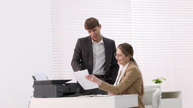Smiling woman holding sheet of paper and talking with her colleague at white desk with printer in office