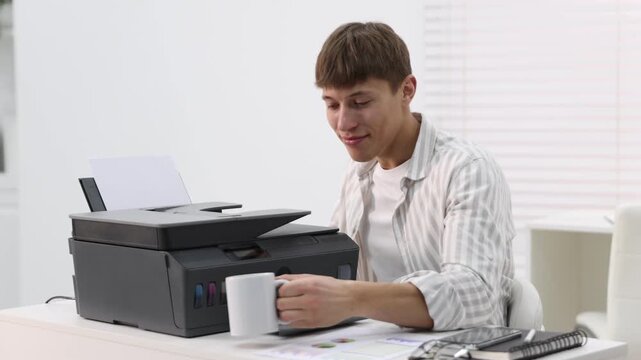 Smiling man with mug loading paper into printer at white desk in office